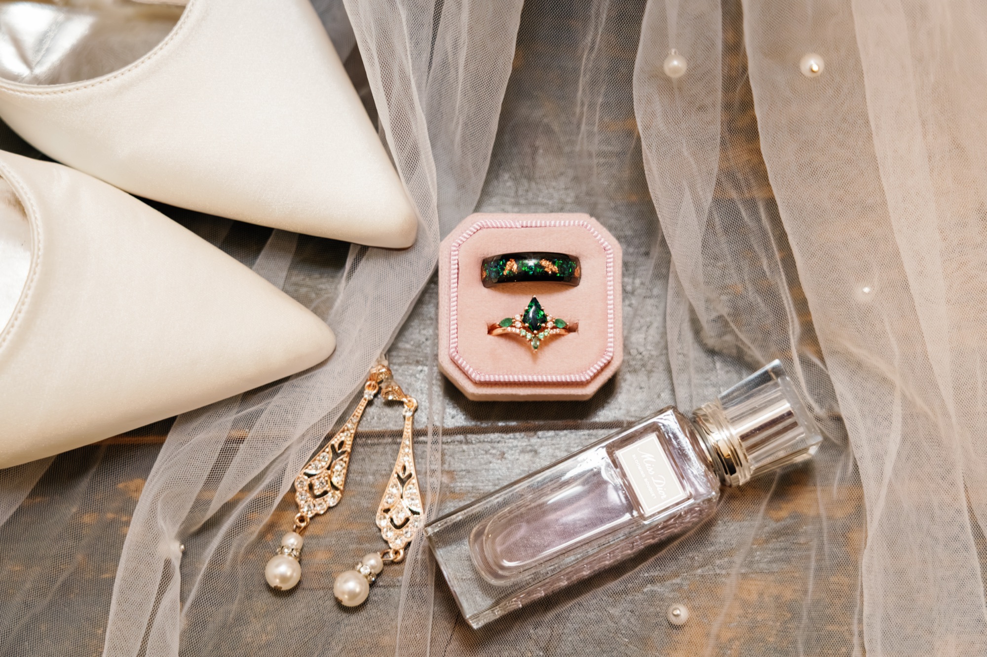 Bridal details flat-lay — an emerald opal ring set in a blush ring box, ivory heels, rose-gold pearl earrings and perfume on a tulle veil, photographed by GM Productions at Kelly and Taylor's wedding
