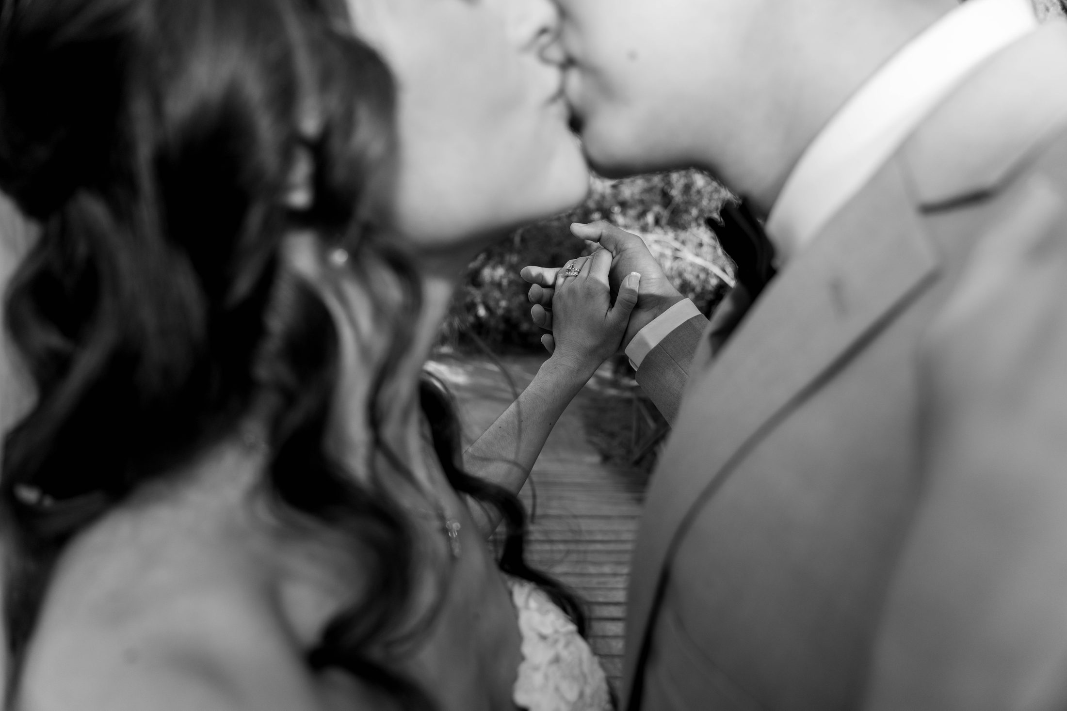 A couple kiss while holding hands, rings visible, in warm black and white